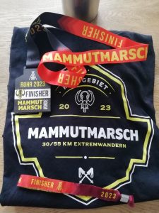 Schwarzes T-Shirt mit der Aufschrift Mammutmarsch Ruhrgebiet 2023, 30/55 Km Extremwandern.
Darauf liegt eine schwarze Holz-Medaille mit rotem Band sowie ein rotes Armband aus Stoff.
Auf beidem steht mit gelber Schrift Finisher.