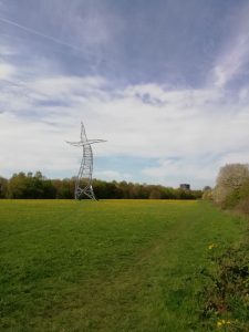 Ein Strommast, der aussieht, als würde er tanzen, auf einer Wiese, im Hintergrund der Gasometer Oberhausen.
Das Kunstwerk heißt Zauberlehrling und steht in Oberhausen.