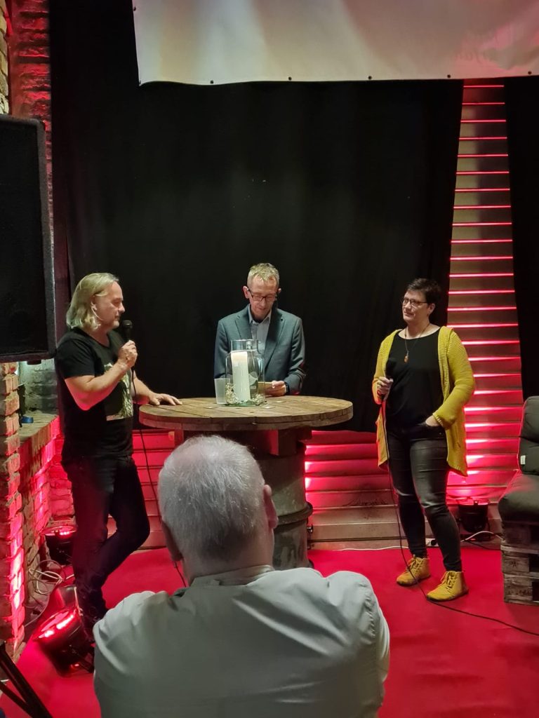 Westerwälder Krimifestival:
Moderator Markus Büth auf der Bühne mit Steffen Ziegler und Melanie Lahmer.
Die Bühne ist rot beleuchtet, alles sieht nach "Industrial-Style" aus.