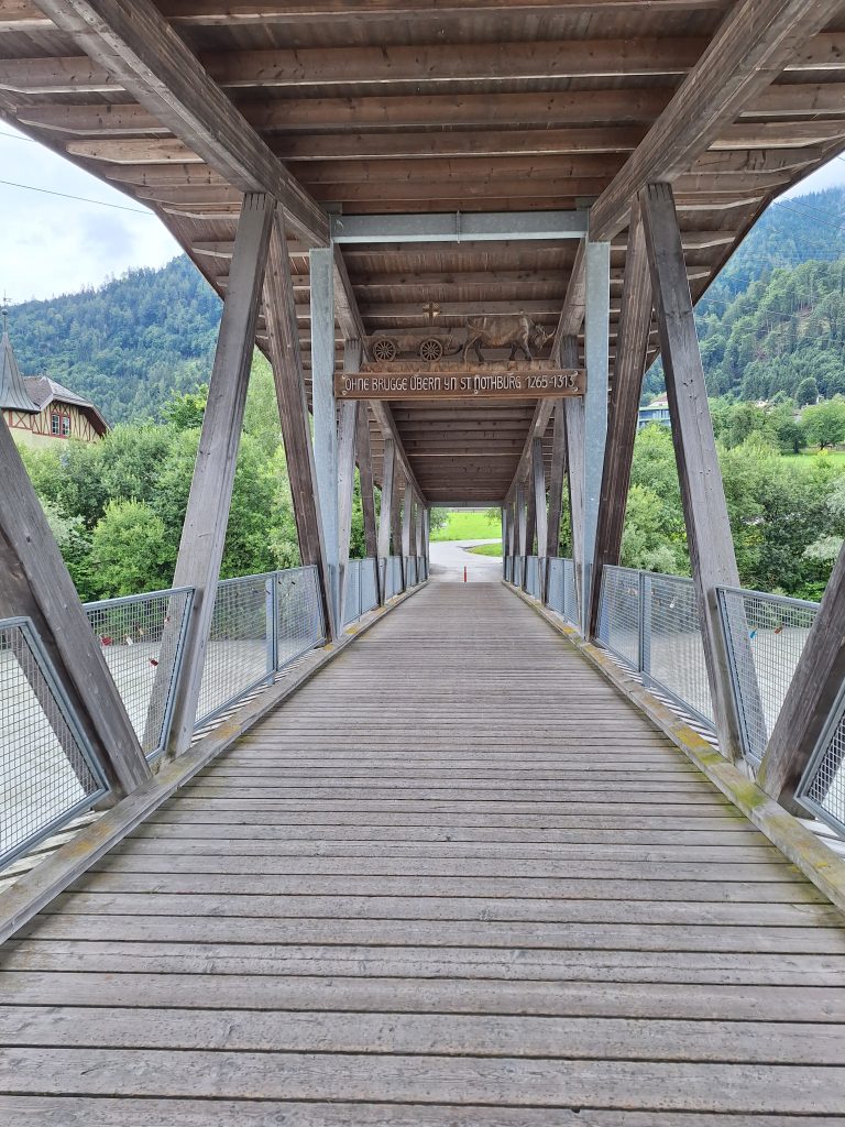 Eine Holzbrücke mit einem Holzdach führt über einen Fluss. In der Mitte der Brücke hängt ein Holzschild mit der Aufschrift "Ohne Brugge übern Yn St. Nothburg 1265-1313". Darüber eine Schnitzerei mit einem Wagen, der von einem Ochsen gezogen wird.