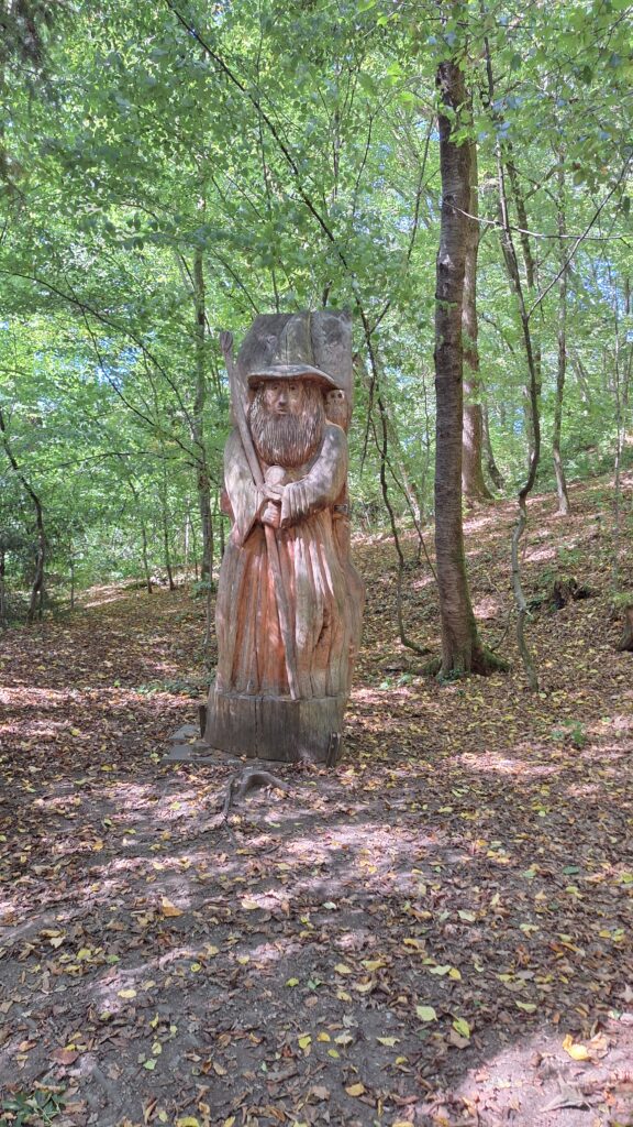Eine mehr als mannshohe geschnitzte Holzfigur - der Zauberer Gandalf aus dem Herrn der Ringe mit Zauberstab auf einer Waldlichtung.
