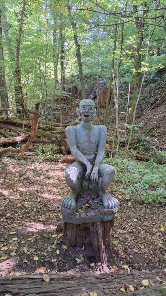 Eine große geschnitzte Holzfigur, Gollum aus dem Herrn der Ringe, sitzt auf einem niedrigen Stamm mitten im Wald.