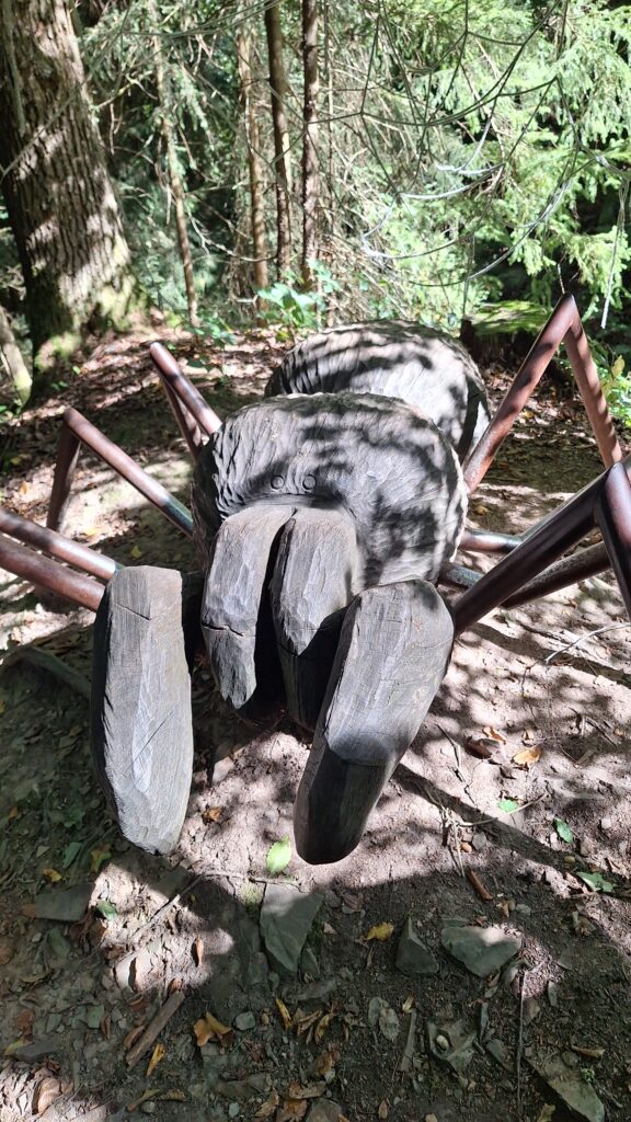 Eine große aus Holz geschnitzte Spinne. Ihre Beine sind aus Metall und sie steht mitten im Wald. Ihre Klauen stehen weit hervor.