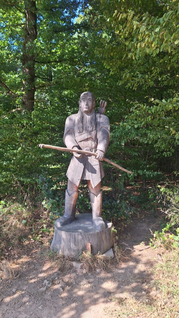 Eine mannshohe geschnitzte Holzfigur - der Elb Legolas aus dem Herrn der Ringe mit Bogen, am Waldrand.